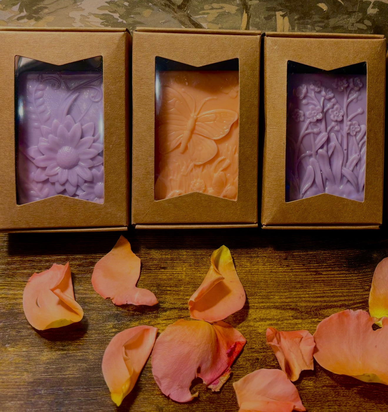 Three floral soap bars in brown boxes with flower petals on a wooden surface